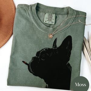 May include: A moss green t-shirt featuring a black graphic of a French Bulldog with a lit cigarette. The shirt has a Comfort Colors tag and a small gold necklace. The word "Moss" is printed on the bottom right.