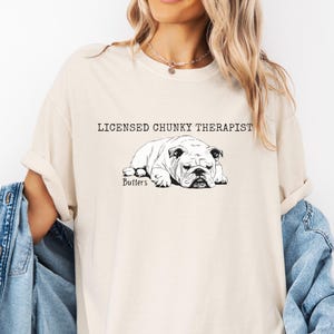 May include: A cream-coloured t-shirt with a black and white bulldog illustration. The text "LICENSED CHUNKY THERAPIST" is above the dog, with "Butters" below. The sleeves are rolled up.