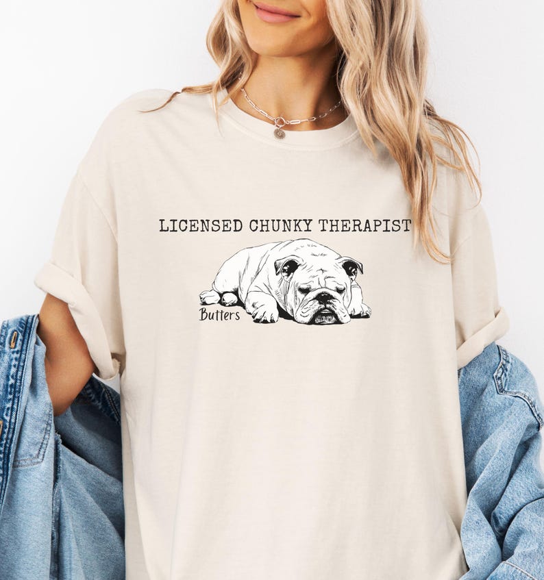 May include: A cream-coloured t-shirt with a black and white bulldog illustration. The text "LICENSED CHUNKY THERAPIST" is above the dog, with "Butters" below. The sleeves are rolled up.