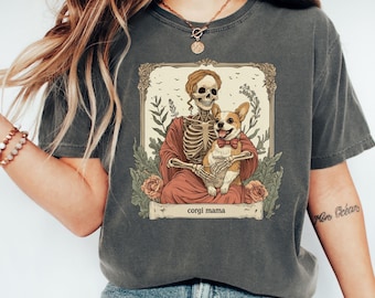 May include: Dark gray t-shirt featuring a skeleton holding a corgi dog wearing a bow tie, with the text "corgi mama" on a banner. The graphic is framed with floral and botanical elements.