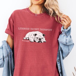 May include: A maroon t-shirt featuring the text "LICENSED CHUNKY THERAPIST" above a black and white bulldog illustration. The bulldog illustration is labeled "Mr. Grumps". The shirt has rolled-up sleeves and a casual fit.