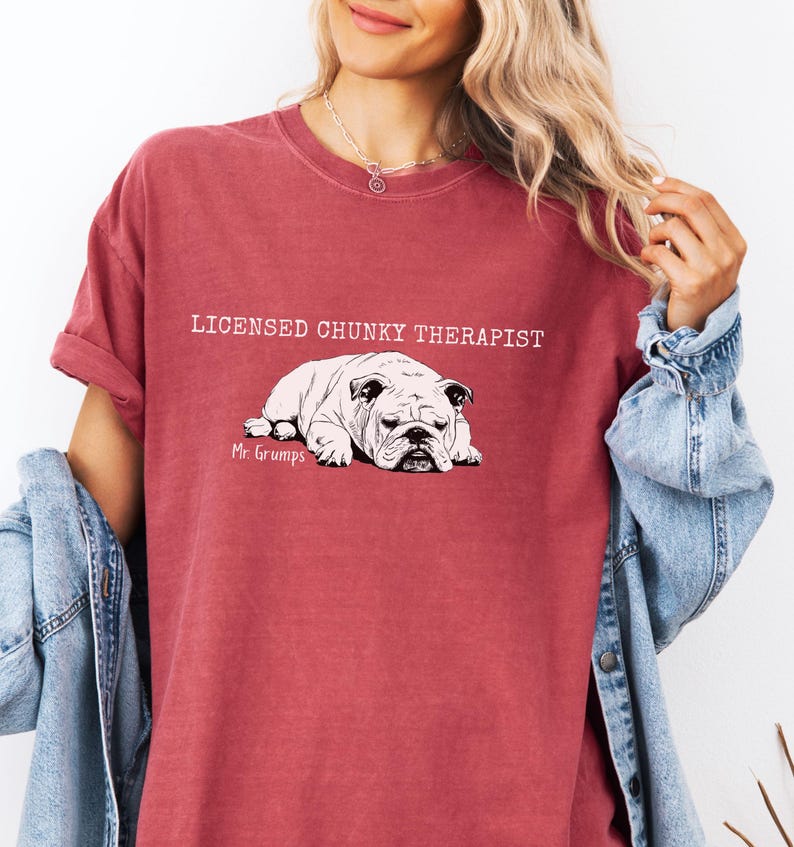 May include: A maroon t-shirt featuring the text "LICENSED CHUNKY THERAPIST" above a black and white bulldog illustration. The bulldog illustration is labeled "Mr. Grumps". The shirt has rolled-up sleeves and a casual fit.