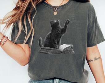May include: Dark gray t-shirt featuring a black cat illustration. The cat is sitting on a stack of books with its paw raised. The graphic is in a vintage style, creating a unique and eye-catching design. This tee is perfect for cat lovers.