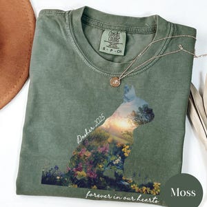 May include: Moss green t-shirt with a cat silhouette filled with a landscape of flowers and trees. The text "Dasher 2025" and "forever in our hearts" are printed on the shirt. A gold necklace with a small pendant rests on the shirt.