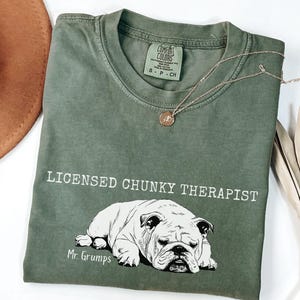 May include: An olive green t-shirt with the words "LICENSED CHUNKY THERAPIST" and a bulldog graphic. The text "Mr. Grumps" is below the bulldog. A gold necklace with a pendant is draped across the shirt. The shirt is folded.