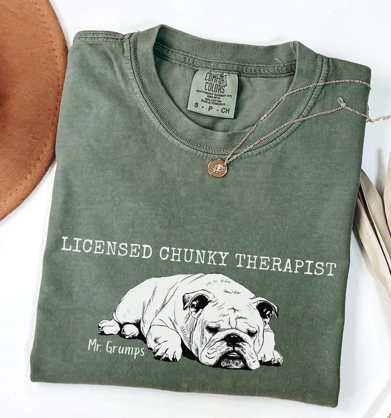 May include: An olive green t-shirt with the words "LICENSED CHUNKY THERAPIST" and a bulldog graphic. The text "Mr. Grumps" is below the bulldog. A gold necklace with a pendant is draped across the shirt. The shirt is folded.