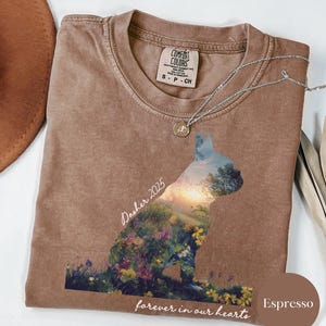 May include: Espresso-colored t-shirt with a dog silhouette filled with a landscape of flowers and a sunset. The shirt has the text "Dasher 2025" and "forever in our hearts". A silver necklace with a pendant is on the shirt.