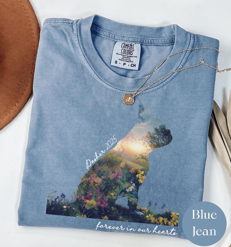 May include: A light blue Comfort Colors t-shirt featuring a dog silhouette filled with a floral landscape and the text "Dasher 2025" and "forever in our hearts". A gold necklace with a pendant rests on the shirt.