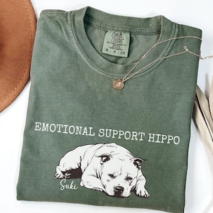 May include: An olive green t-shirt with the text "EMOTIONAL SUPPORT HIPPO" and a graphic of a dog. The dog's name, "Suki", is printed below the graphic. A gold necklace with a pendant is draped across the shirt.