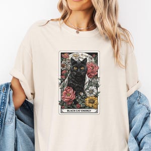 May include: Cream-colored t-shirt featuring a tarot card design with a black cat, surrounded by flowers. The card reads "BLACK CAT ENERGY" in a rectangular frame. The cat has yellow eyes and is set against a floral and moon background.