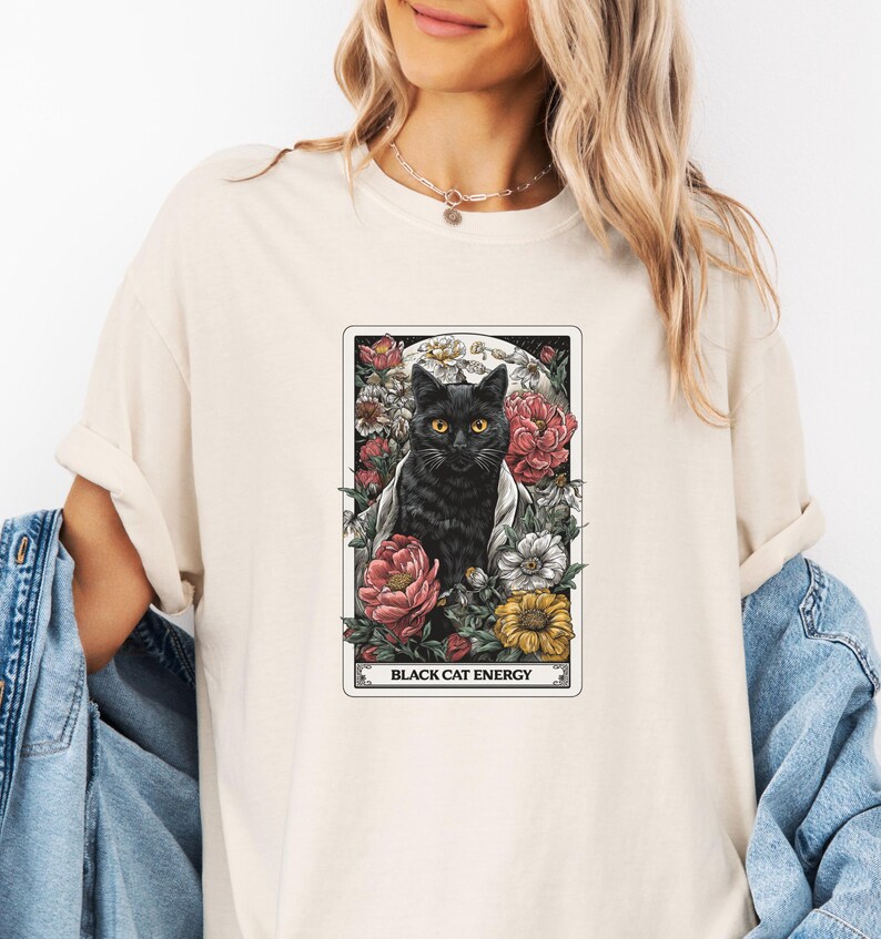 May include: Cream-colored t-shirt featuring a tarot card design with a black cat, surrounded by flowers. The card reads "BLACK CAT ENERGY" in a rectangular frame. The cat has yellow eyes and is set against a floral and moon background.