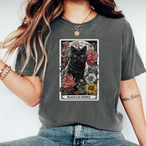 May include: Dark grey t-shirt featuring a tarot card design with a black cat surrounded by flowers. The card reads "Black Cat Energy" in white text. The cat has yellow eyes and is set against a full moon.