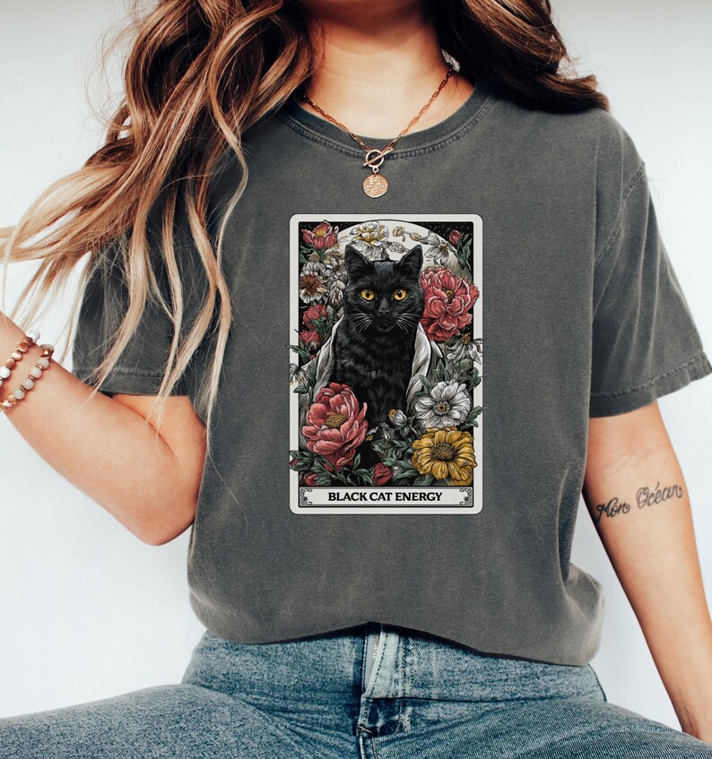 May include: Dark grey t-shirt featuring a tarot card design with a black cat surrounded by flowers. The card reads "Black Cat Energy" in white text. The cat has yellow eyes and is set against a full moon.