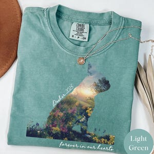 May include: Light green Comfort Colors t-shirt with a dog silhouette filled with a floral landscape. The text "Dasher 2025" and "forever in our hearts" are printed on the shirt. A silver necklace with a pendant is draped on the shirt.