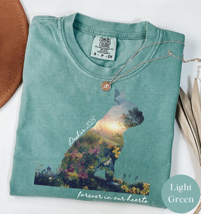 May include: Light green Comfort Colors t-shirt with a dog silhouette filled with a floral landscape. The text "Dasher 2025" and "forever in our hearts" are printed on the shirt. A silver necklace with a pendant is draped on the shirt.