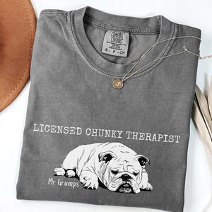 May include: A grey t-shirt featuring the words "LICENSED CHUNKY THERAPIST" above a black and white bulldog illustration. The bulldog is labelled "Mr. Grumps". A silver necklace with a circular pendant is draped across the shirt.