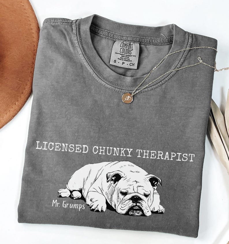 May include: A grey t-shirt featuring the words "LICENSED CHUNKY THERAPIST" above a black and white bulldog illustration. The bulldog is labelled "Mr. Grumps". A silver necklace with a circular pendant is draped across the shirt.