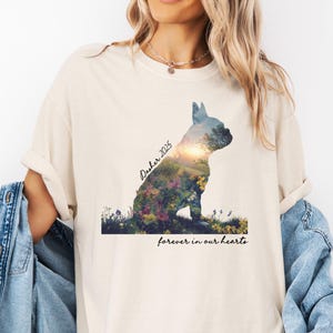 May include: Cream-colored t-shirt featuring a silhouette of a dog filled with a floral landscape. The text "Dasher 2025" is above the dog, and "forever in our hearts" is below. The shirt has rolled-up sleeves.