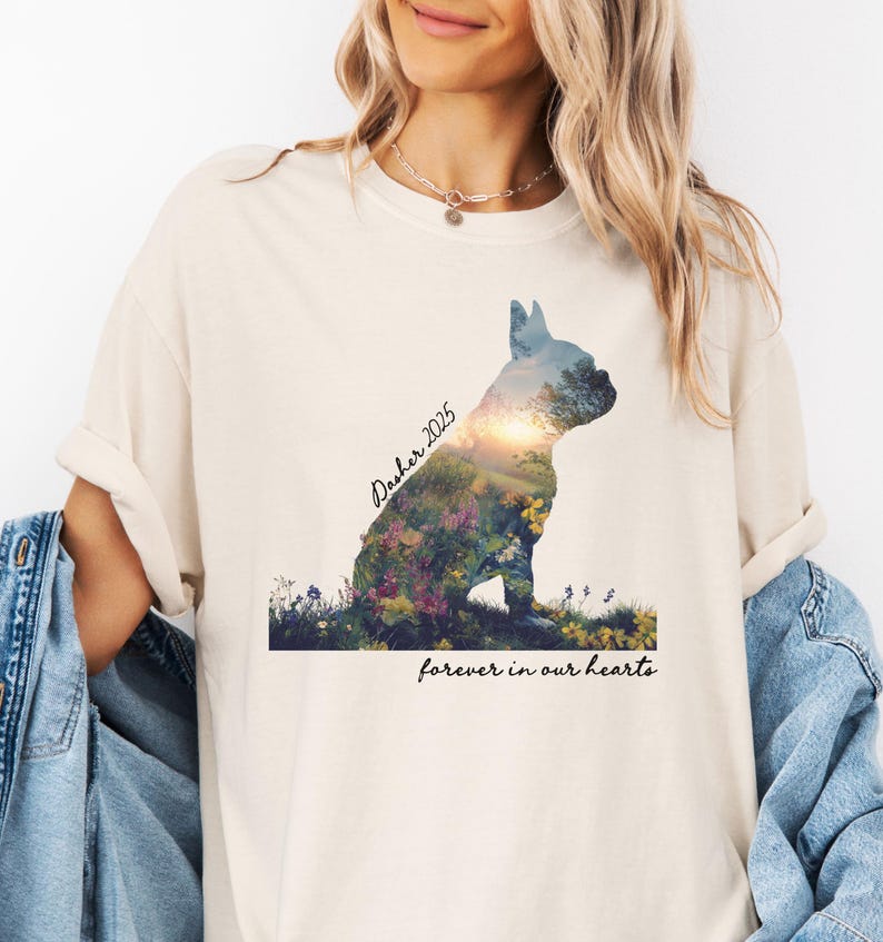 May include: Cream-colored t-shirt featuring a silhouette of a dog filled with a floral landscape. The text "Dasher 2025" is above the dog, and "forever in our hearts" is below. The shirt has rolled-up sleeves.