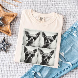 Pu&ograve; includere: T-shirt color crema con un'immagine in bianco e nero a quattro pannelli di un chihuahua con occhiali da sole. Il cane tiene anche una sigaretta in due dei pannelli. La maglietta &egrave; piegata sopra jeans blu strappati.
