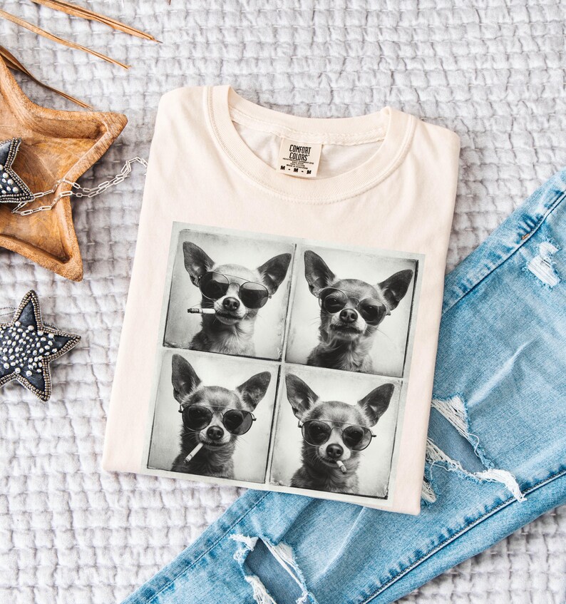 Pu&ograve; includere: T-shirt color crema con un'immagine in bianco e nero a quattro pannelli di un chihuahua con occhiali da sole. Il cane tiene anche una sigaretta in due dei pannelli. La maglietta &egrave; piegata sopra jeans blu strappati.