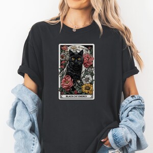 May include: Black t-shirt featuring a tarot card design with a black cat surrounded by flowers. The card reads "BLACK CAT ENERGY" in white text. The cat has yellow eyes and is set against a moonlit background.