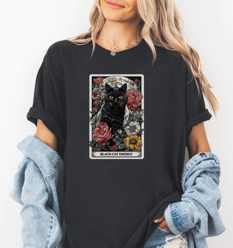 May include: Black t-shirt featuring a tarot card design with a black cat surrounded by flowers. The card reads "BLACK CAT ENERGY" in white text. The cat has yellow eyes and is set against a moonlit background.
