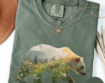 May include: A moss green t-shirt featuring a bear silhouette filled with a landscape of flowers and mountains. The shirt has a crew neck and short sleeves. A silver necklace with a small pendant rests on the shirt.