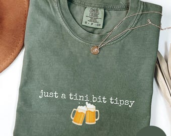 May include: A moss green t-shirt with the phrase "just a tini bit tipsy" and two beer mugs printed on the front. A silver necklace with a small pendant is draped across the shirt. The shirt is folded.