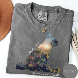 May include: A gray t-shirt with a cat silhouette filled with a colorful landscape of flowers and trees. The text "Dasher 2025" and "forever in our hearts" are printed on the shirt. A silver necklace with a pendant is draped on the shirt.
