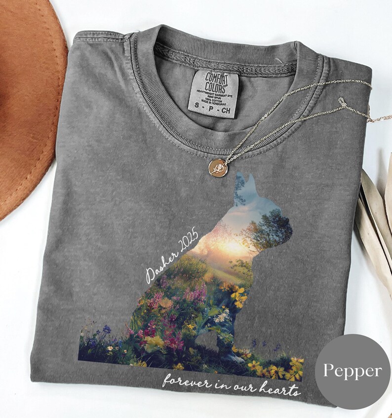 May include: A gray t-shirt with a cat silhouette filled with a colorful landscape of flowers and trees. The text "Dasher 2025" and "forever in our hearts" are printed on the shirt. A silver necklace with a pendant is draped on the shirt.