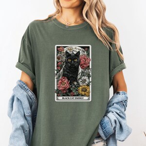 May include: Olive green t-shirt featuring a tarot card design with a black cat surrounded by flowers and a full moon. The text "BLACK CAT ENERGY" is displayed at the bottom of the card.