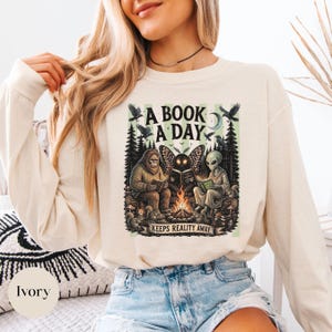 May include: An ivory long-sleeve sweatshirt featuring a graphic print of a Bigfoot, a moth-like creature, and an alien sitting around a campfire reading books. The text reads A BOOK A DAY KEEPS REALITY AWAY, surrounded by trees, birds, and squirrels.