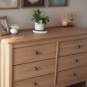May include: A six-drawer dresser made of light wood with brass drawer pulls. The dresser has a light wood top and is sitting on a light wood floor. There are three framed pictures on the wall behind the dresser.