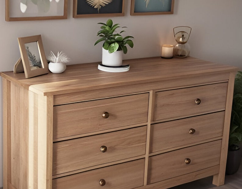 May include: A six-drawer dresser made of light wood with brass drawer pulls. The dresser has a light wood top and is sitting on a light wood floor. There are three framed pictures on the wall behind the dresser.