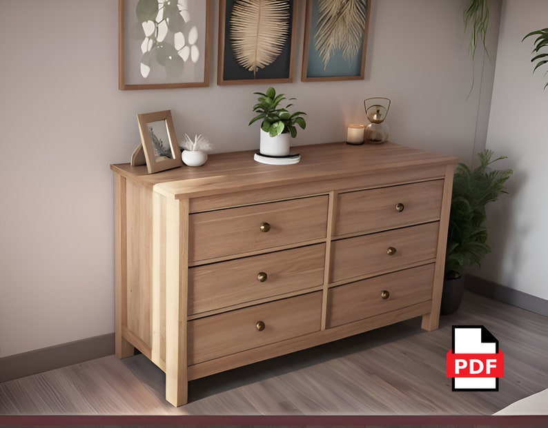 May include: A light wood dresser with six drawers and brass knobs. The dresser is in a room with a light wood floor and a white wall. There are framed pictures and plants on the wall above the dresser.