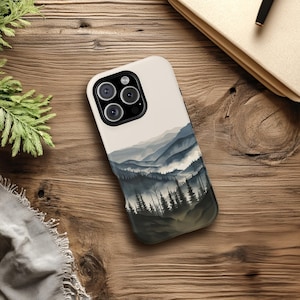 May include: A white phone case with a watercolor design of a mountain range with fog and trees. The case has a black camera lens cutout.