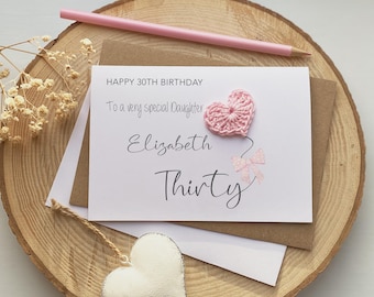 May include: A birthday card with a crocheted pink heart and bow, reading "Happy 30th Birthday" and "To a very special Daughter Elizabeth Thirty". A pink pencil lies on a wooden slice. The card is for a daughter.