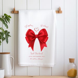 May include: White kitchen towel featuring a large red bow design. The towel is printed with the names "Stephen" and "Rachael", the text "Tied the knot" and a date. A heart graphic is between the names.