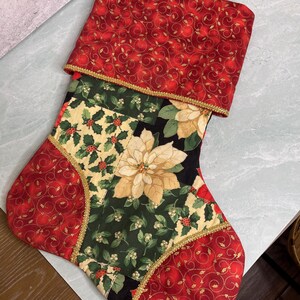 May include: A Christmas stocking featuring a red cuff with gold swirling patterns and a gold trim. The body is a patchwork of fabrics including red with gold swirls, green with holly berries, and a central panel with large cream poinsettia flowers.