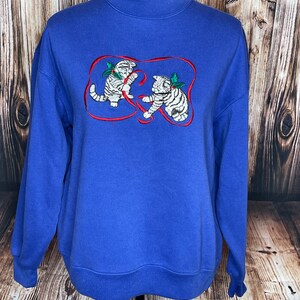 May include: A royal blue long-sleeve sweatshirt featuring an embroidered graphic of two white kittens playing with a red ribbon. Each kitten has a small green holly sprig with red berries attached to its head. The sweatshirt has a mock neck collar.