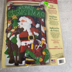 May include: A Christmas felt applique kit by Dimensions. The kit includes a banner with a Santa Claus design, a Christmas tree, and reindeer. The banner reads "MERRY CHRISTMAS" and measures approximately 15" x 22".