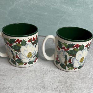 May include: Two ceramic mugs with a white exterior and a dark green interior. The mugs feature a botanical pattern of white magnolia flowers, green leaves, and clusters of red holly berries wrapping around the outer surface of each vessel.