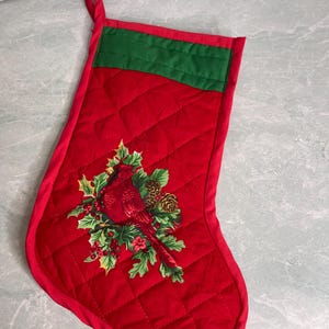 May include: A quilted red Christmas stocking featuring a green fabric cuff at the top and a hanging loop. The center of the stocking displays a detailed applique of a red cardinal bird perched among green holly leaves, red berries, and pinecones.