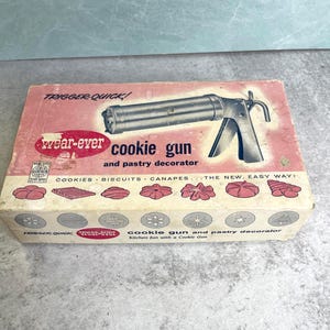 May include: A rectangular cardboard box for a Wear-Ever Trigger-Quick cookie gun and pastry decorator. The box features a pink and white design with an illustration of the metal tool, text, and various cookie shapes printed on the front.