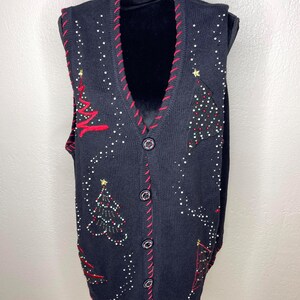 May include: A black knit sweater vest featuring a V-neckline with red stitched trim. The vest is decorated with embroidered Christmas tree motifs, small gold star accents, and scattered white beads. It has a button-down front with five dark buttons.