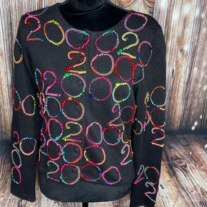 May include: Black cardigan sweater with colorful beaded accents. The sweater features the number "2022" in various places, outlined with vibrant beads in shades of red, pink, yellow, green, and blue. The sweater has a round neckline and button closure.