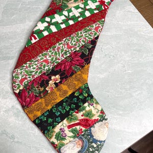 May include: A Christmas stocking featuring a diagonal patchwork design of various holiday-themed fabrics, including poinsettias, holly, cardinals, and Santa Claus. The top cuff is made of red fabric with a small white polka dot pattern.