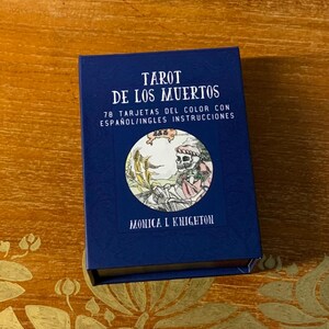 Pode incluir: Uma caixa azul com o t&iacute;tulo "Tarot de los Muertos" e o texto "78 Tarjetas del Color con Espa&ntilde;ol/Ingles Instrucciones". A caixa apresenta uma imagem circular de um esqueleto segurando uma foice em um campo de trigo. O texto "Monica L Knighton" est&aacute; impresso abaixo da imagem.