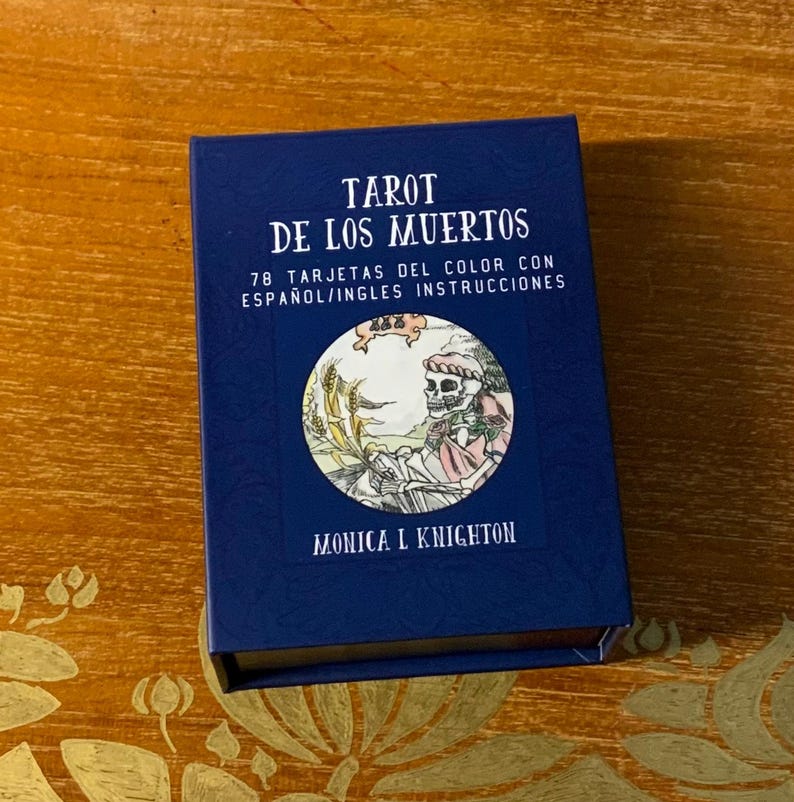 Pode incluir: Uma caixa azul com o t&iacute;tulo "Tarot de los Muertos" e o texto "78 Tarjetas del Color con Espa&ntilde;ol/Ingles Instrucciones". A caixa apresenta uma imagem circular de um esqueleto segurando uma foice em um campo de trigo. O texto "Monica L Knighton" est&aacute; impresso abaixo da imagem.